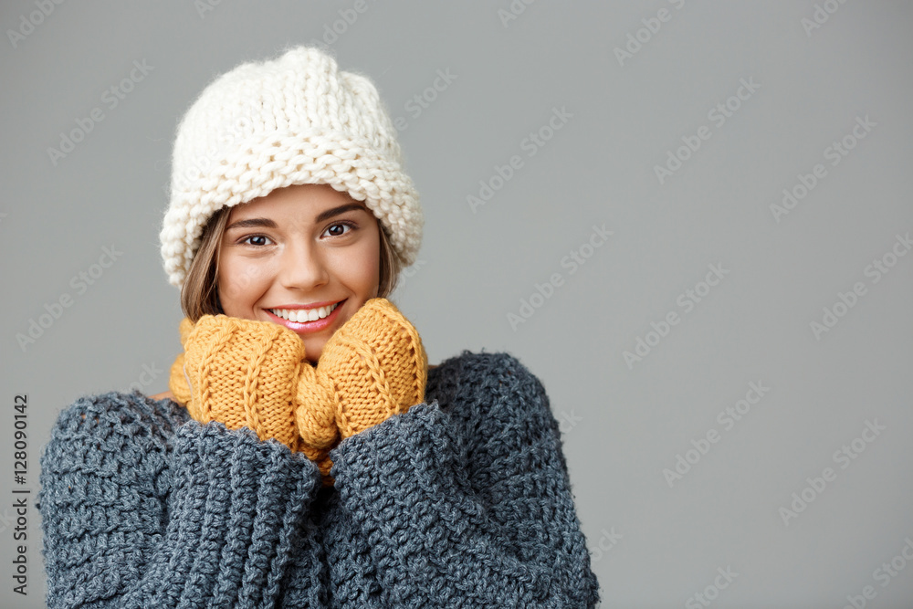 Young beautiful fair-haired girl in knited hat sweater scarf and mittens smiling looking at camera over grey background. Young beautiful fair-haired girl in knited hat sweater scarf and mittens smiling looking at camera over grey background.
