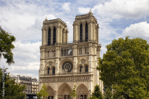 View of Notre Dame Cathedral with trees and cloudy sky in Paris. Towering, 13th-century cathedral with flying buttresses & gargoyles, setting for Hugo's novel.