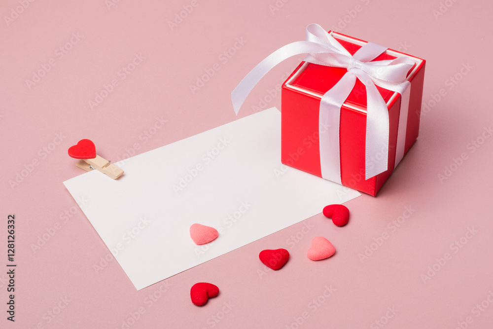 Valentine day composition: red gift box with bow, stationery / photo template with clamp and small hearts on light pink background. Valentine day composition: red gift box with bow, stationery / photo template with clamp and small hearts on light pink background.