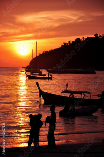 a silhouette of family standing around seashore with boats and sunset sky background