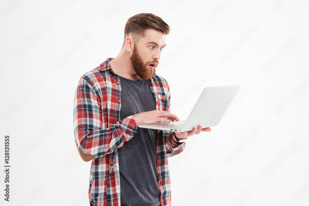 Surprised young bearded man in plaid shirt looking at laptop Surprised young bearded man in plaid shirt looking at laptop