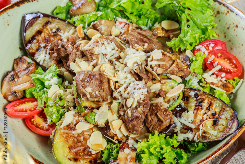 Hot salad with veal, mushrooms, salad leaves, eggplant, zucchini, tomatoes, garnished with grated almonds and Parmesan cheese and glass of wine on wooden background. Healthy food Hot salad with veal, mushrooms, salad leaves, eggplant, zucchini, tomatoes, garnished with grated almonds and Parmesan cheese and glass of wine on wooden background. Healthy food