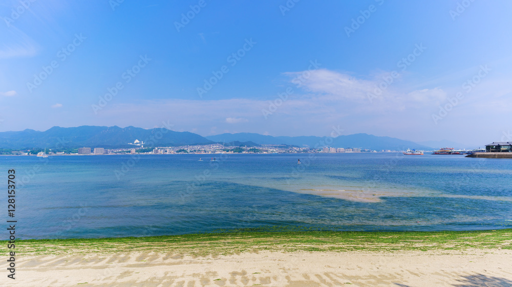 Fototapeta premium Scenery of sea coast on Miyajima Island where Itsukushima shrine located viewing ferry terminal , activities in the bay and Hiroshima city in the background , Japan