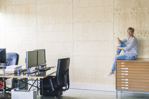 Smiling businesswoman sitting on filing cabinet in creative office