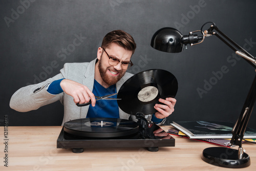 Mad irritated man sitting and cutting vinyl record with scissors