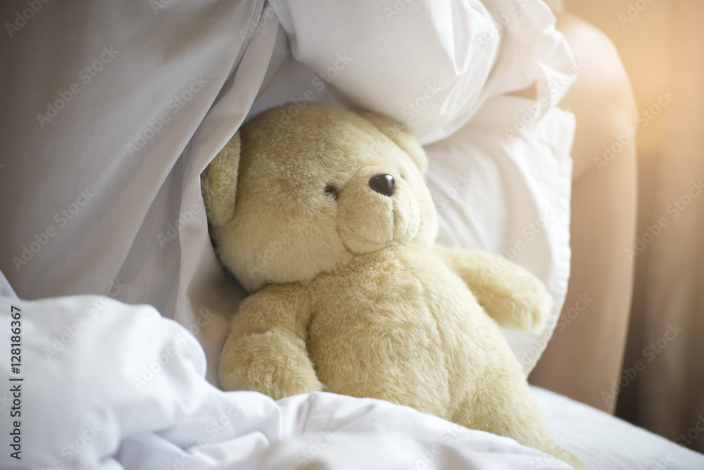 Beautiful woman with long hair wearing white pajamas,sitting on a white bed with teddy bear,in the morning.