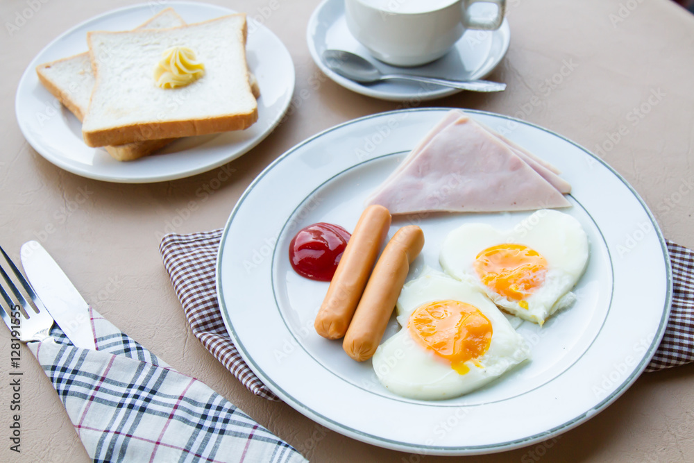 Breakfast with fried egg, ham, sausage, toast, butter, and coffee on table.