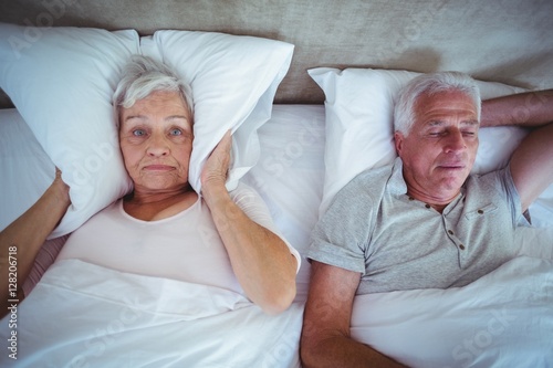 Wife blocking ears with pillow while husband snoring on bed