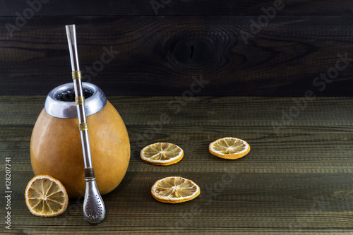 Yerba mate in gourd matero with bombila and dried lemon slices on wooden background.