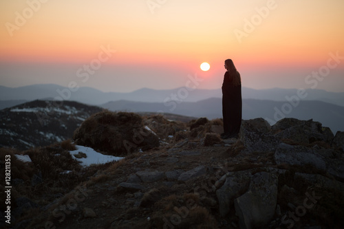 Young woman in long mystical dress looking into the sun stands on the top of the mountain. Out of focus