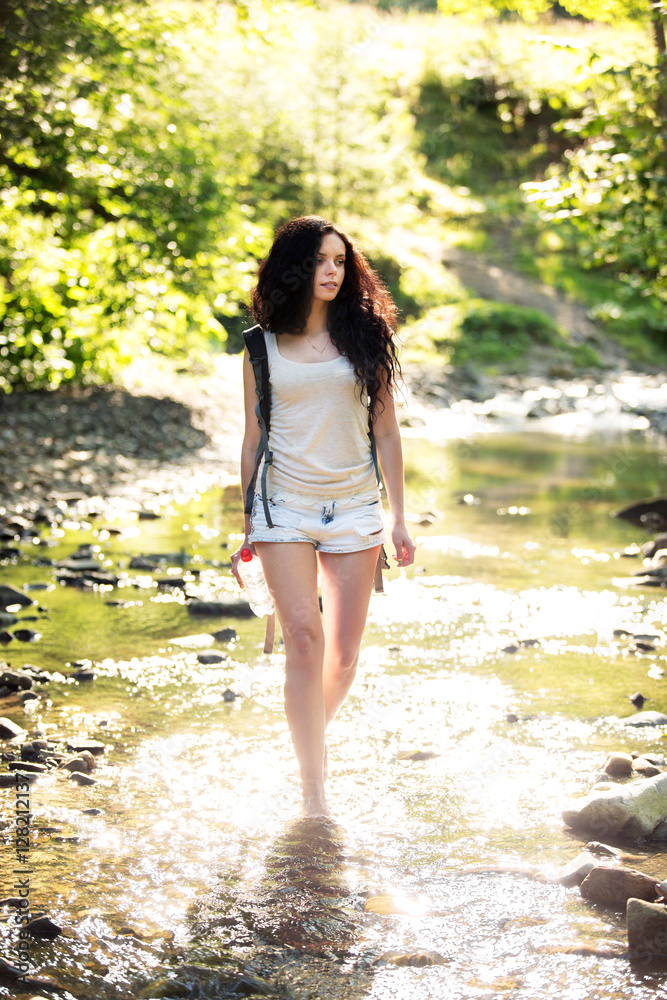 Outdoor shot of attractive young woman with backpack standing in a mountain stream. Female hiker in creek water.Portrait of young pretty brunette woman holding bottle of water. Outdoor shot of attractive young woman with backpack standing in a mountain stream. Female hiker in creek water.Portrait of young pretty brunette woman holding bottle of water.