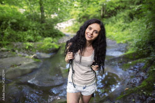 Portrait of beautiful young woman with backpack standing by the creek. Female hiker hiking by the mountain stream.