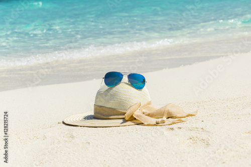 White female trendy hat with blue aviator sunglasses lying on white sand beach in front of turquoise ocean.