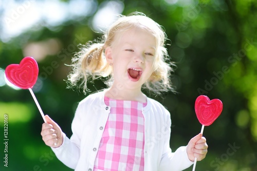 Cute little girl eating huge heart-shaped lollipops outdoors