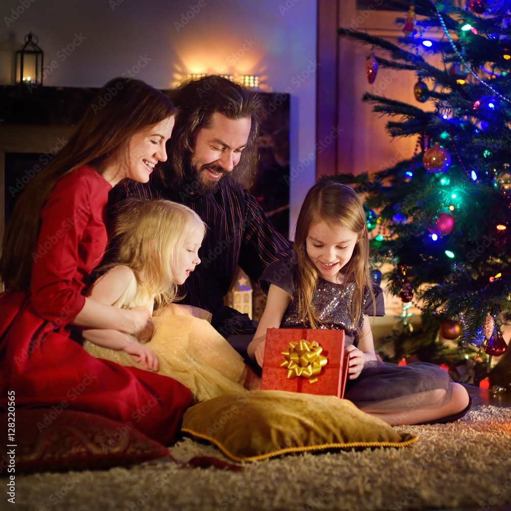 Young happy family of four unwrapping Christmas gifts by a fireplace