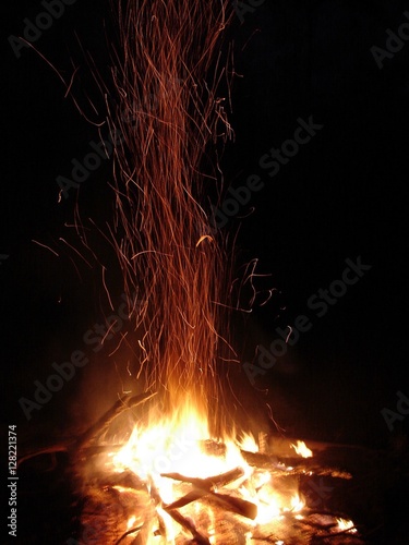 Sparks fly off a burning campfire against a black night sky.