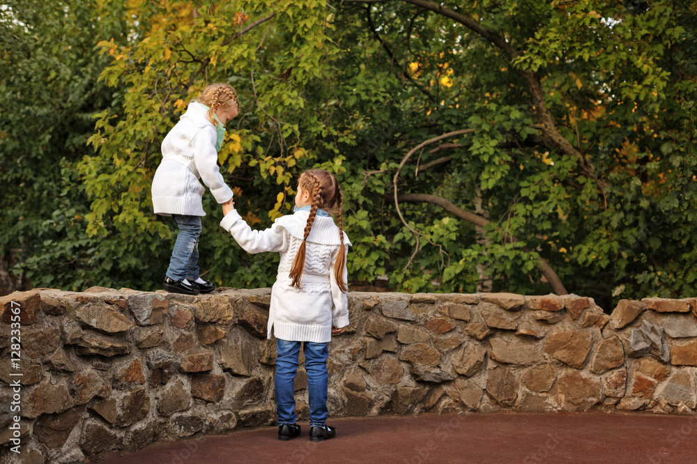 Fototapeta premium Cute little sisters in the autumn park. The older girl helping younger go on the stone fence. Family time. Sport games.