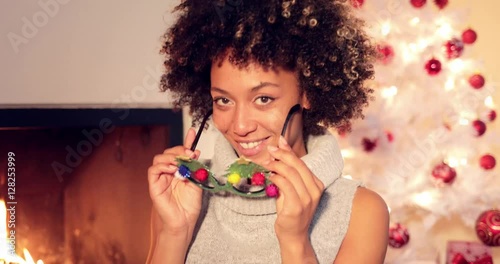 Fun vivacious young woman celebrating Christmas wearing a colorful pair of party goggles laughing at the camera in front of a burning fire and decorated tree. photo