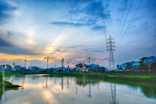 Sunset in urban areas, featuring transmission towers, ponds that make beautiful reflection, crane, rays of light and many houses