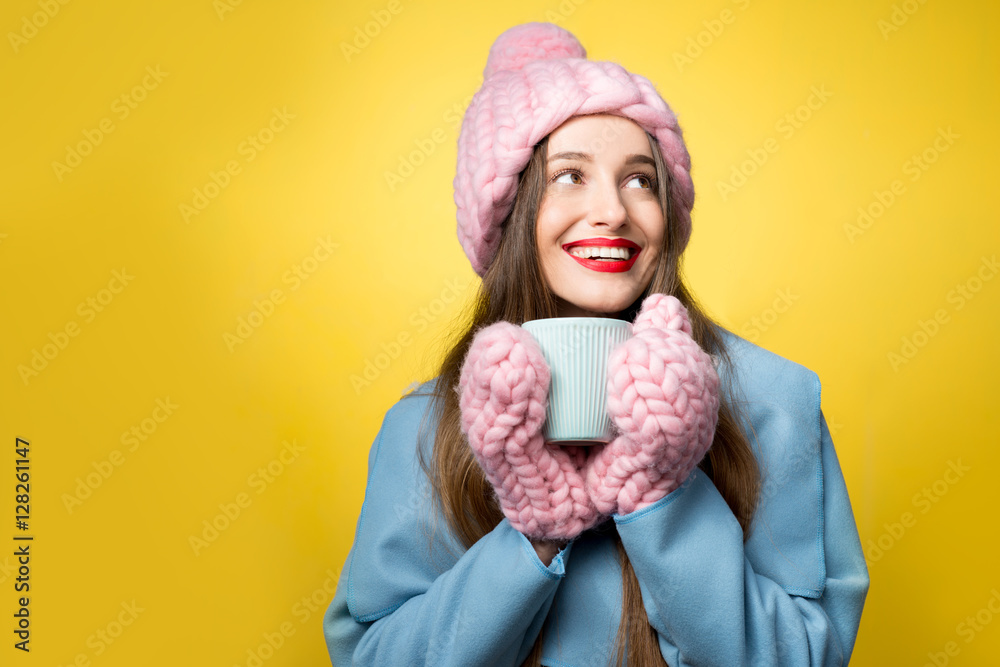 Woman in colorful winter clothes holding a cup of hot drink standing on the yellow background. Happy winter concept