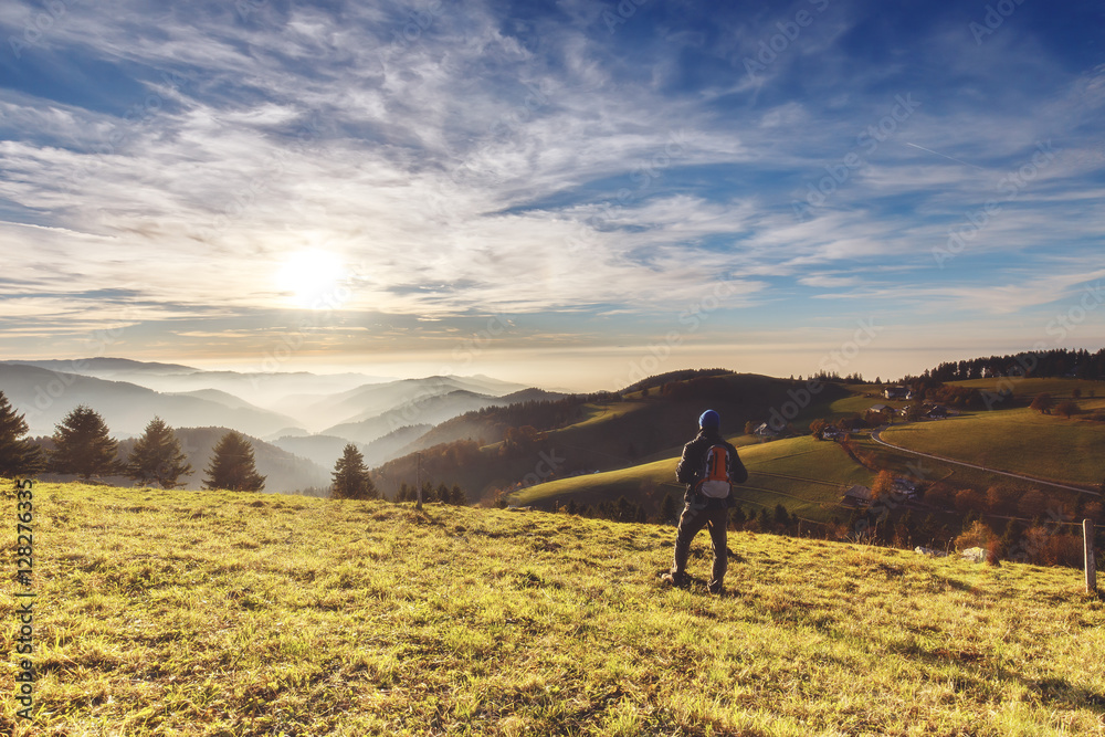 Man backpacker hiking in mountains in autumn looking at beautiful landscape with mountains forest and a village at sunset.