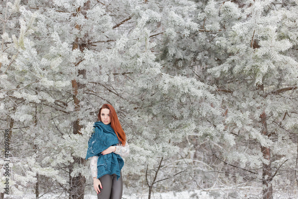 beautiful red-haired girl with a scarf on his shoulders walking in the winter forest of firs and pines stilled