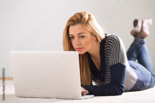Beautiful adult woman lying on floor at home and using laptop