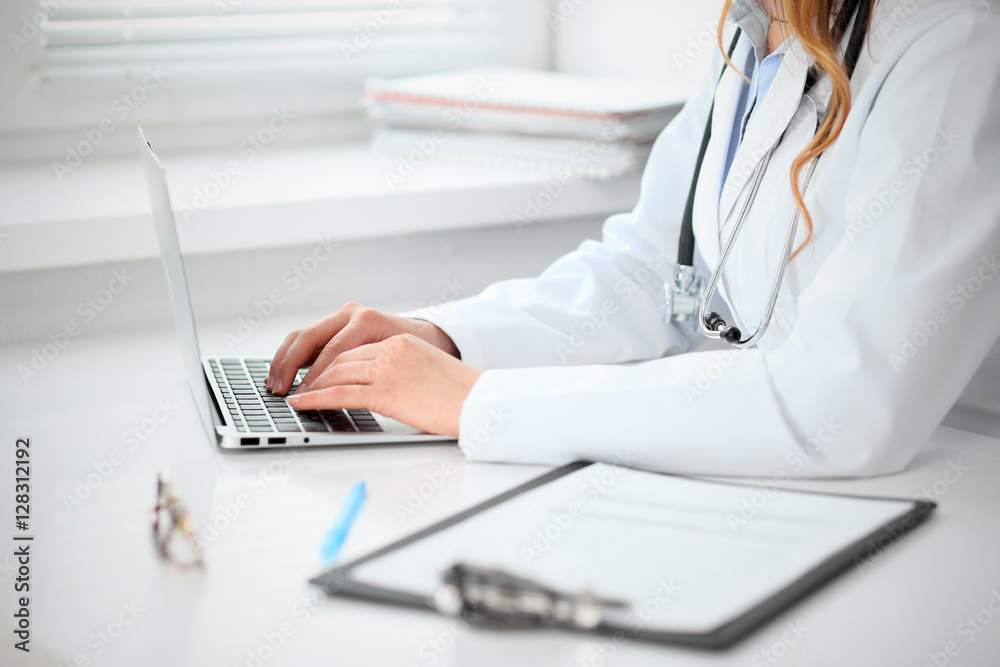 Close up of  unknown female doctor sitting  at the table near the window in hospital and typing at laptop computer