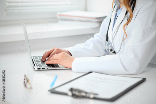Close up of unknown female doctor sitting at the table near the window in hospital and typing at laptop computer