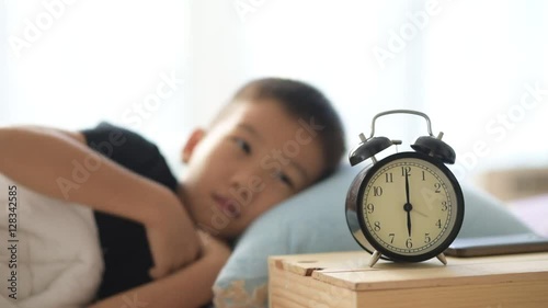 Selective focus at vintage alarm clock show at six o clock and out focus young asian boy sleeping and wake up on the bed.