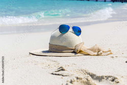 White female trendy hat with blue aviator sunglasses lying on white sand beach in front of turquoise ocean.