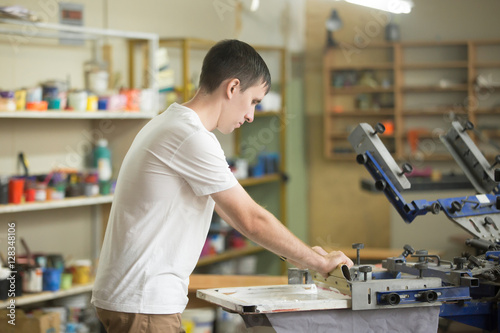 Young confident, experienced male working with squeegee on a small factory, handsome worker man using printmaking tools, screen printing on clothing fabric technique
