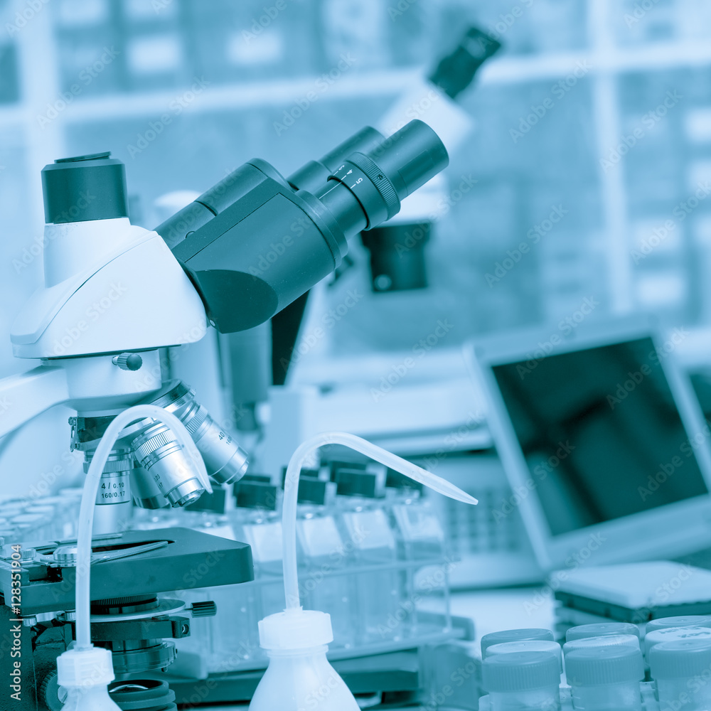 Science laboratory with a microscope, biological material samples and a computer on a desk. Selective focus with bokeh. Rack with samples and chemicals on the background. Blue toned image Science laboratory with a microscope, biological material samples and a computer on a desk. Selective focus with bokeh. Rack with samples and chemicals on the background. Blue toned image