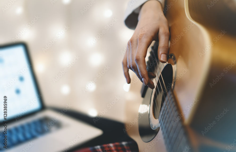 Hipster girl playing guitar in home atmosphere, person studying on musical instrument and notes in laptop on background glow bokeh Christmas illumination, holiday on relax glitter decoration, blur