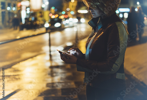 Girl pointing finger on screen smartphone on background illumination glow bokeh light in night christmas city, smiling hipster using in hands mobile phone, headlights taxi; mockup glitter street