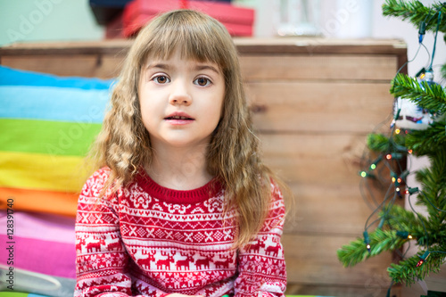 Small Caucasian girl portrait in sweater, sitting on the floor near Christmas tree © Kekyalyaynen