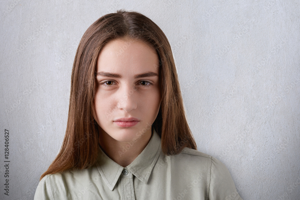Obraz premium A close-up of a beautiful young girl with shining eyes and straight long dark hair having a sombre face looking straight in the camera. A portrait of cute girl on white background falling to thinking