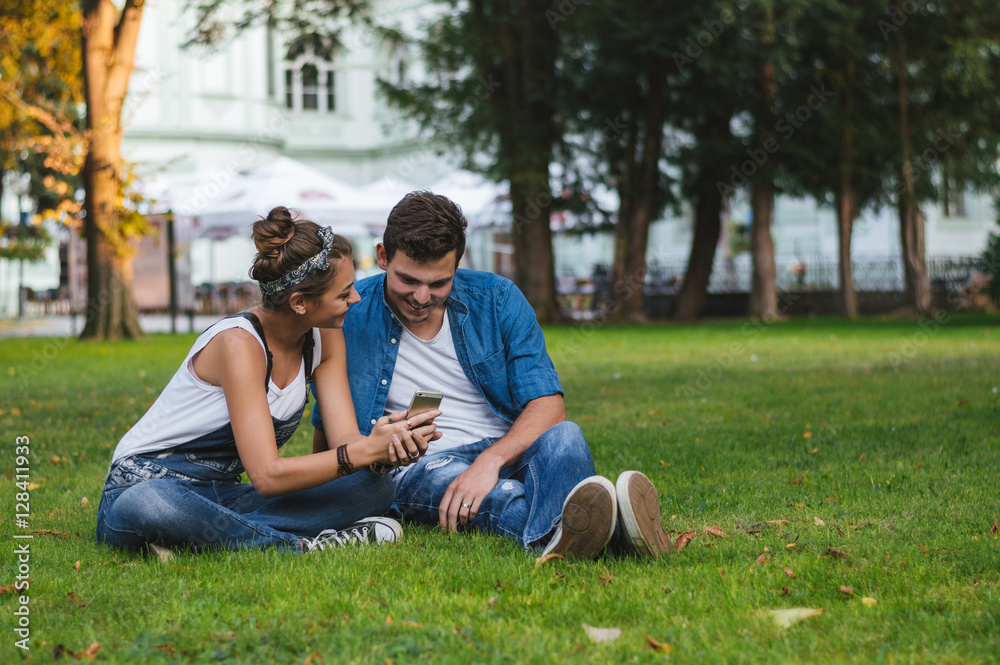 Couple sharing media in a smart phone sitting in a park with bui Couple sharing media in a smart phone sitting in a park with bui