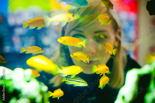 Young blonde woman looking through a window on the life of aquarium fish. aquarium cichlid exotic fish. a flock of beautiful fish swimming in an aquarium.