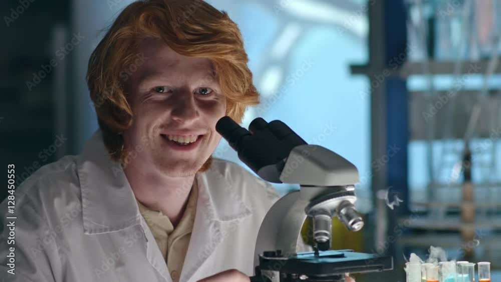 Closeup of young redhead chemistry student looking through microscope and then looking at the camera and smiling Closeup of young redhead chemistry student looking through microscope and then looking at the camera and smiling