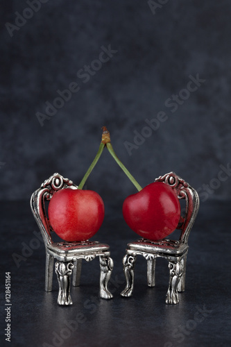 Couple of red cherries placed on beautiful silver vintage chairs on dark background with copy space. Relationship concept, vertical image.