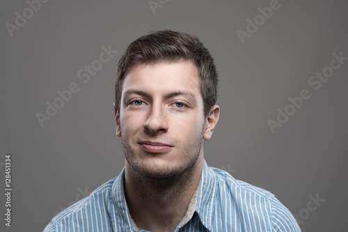 Moody headshot portrait of young man in blue shirt with smirk smile looking at camera photo