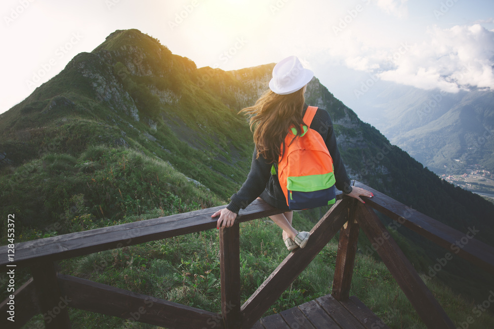 Young traveling woman wearing hat and backpack sitting high on the top of the mountain with waving hair, evening with sunset - obrazy, fototapety, plakaty 