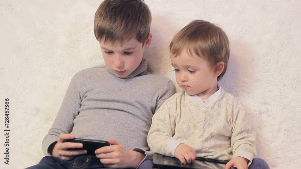 Two boys playing with the tablet and the phone sitting on a white sofa Two boys playing with the tablet and the phone sitting on a white sofa