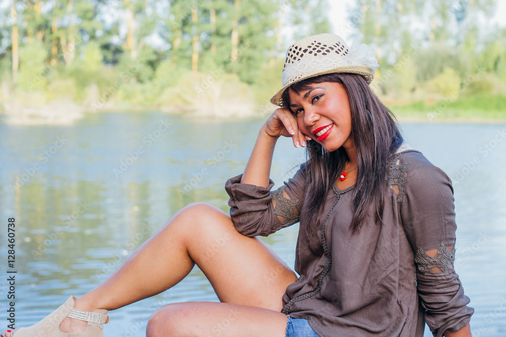 Beautiful black woman posing on a lake. She is wearing a hat and is smiling.