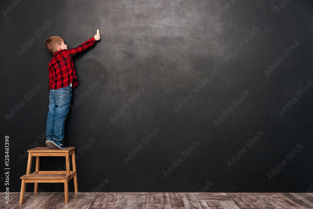 Child standing on stool over chalkboard and drawing at board - obrazy, fototapety, plakaty 