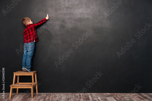 Child standing on stool over chalkboard and drawing at board