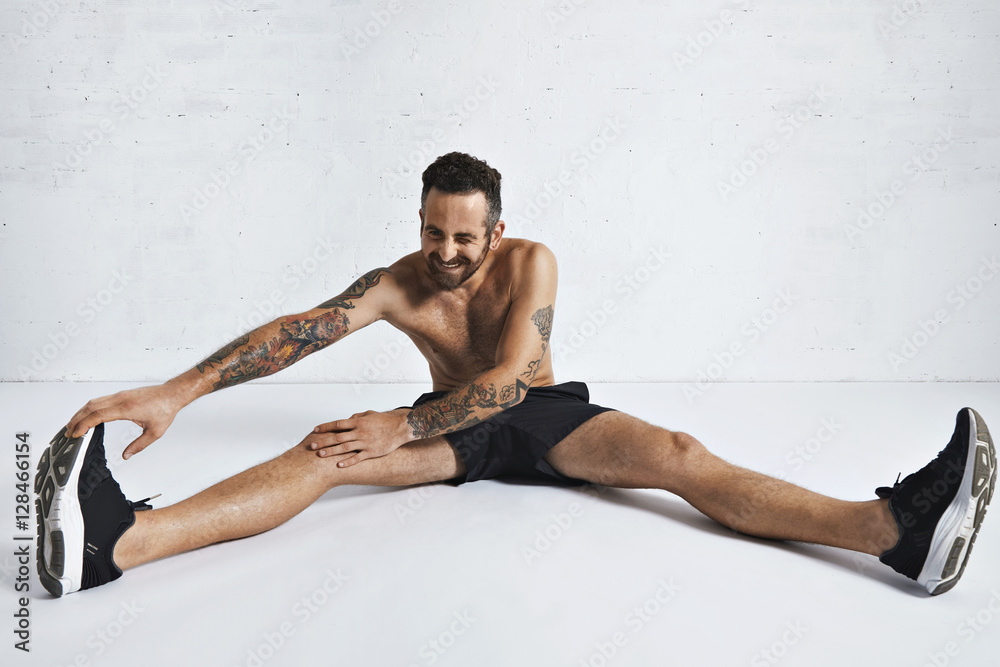 Obraz premium Bearded attractive tattooed male athlete stretches his legs, sitting in white room and smling on camera