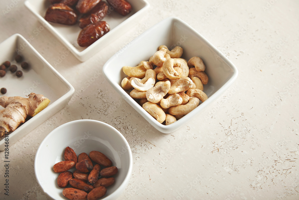 Cashews, brazilian nuts, ginger, pepper and dates arranged in small round and square white porcelain bowls on old white painted table with rust stains