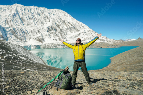 Travel concept: Happy hiker with rised hands, tourist sticks and backpack on Tilicho lake. Its 4900m above sea level. Snowly mountains and Tilicho lake on background. photo